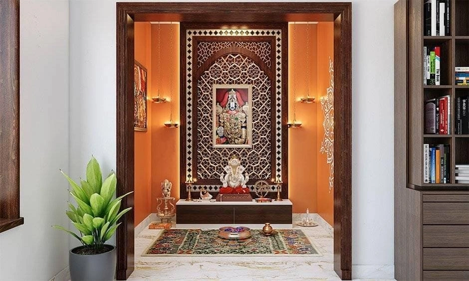 Simple pooja room designs with an orange backdrop wall, hanging brass diyas and a carved marble altar.